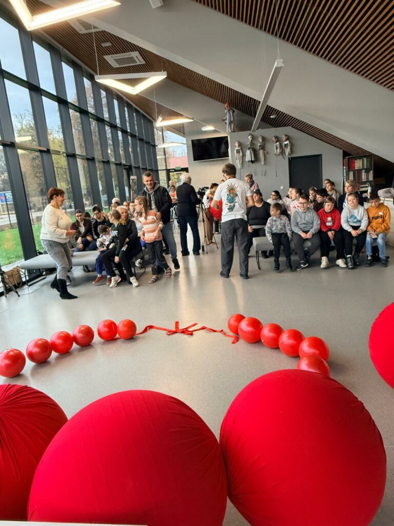 Grupa osób siedzi i stoi w przestronnej sali z dużymi oknami. Na podłodze leżą czerwone piłki, a uczestnicy czekają na rozpoczęcie zajęć.