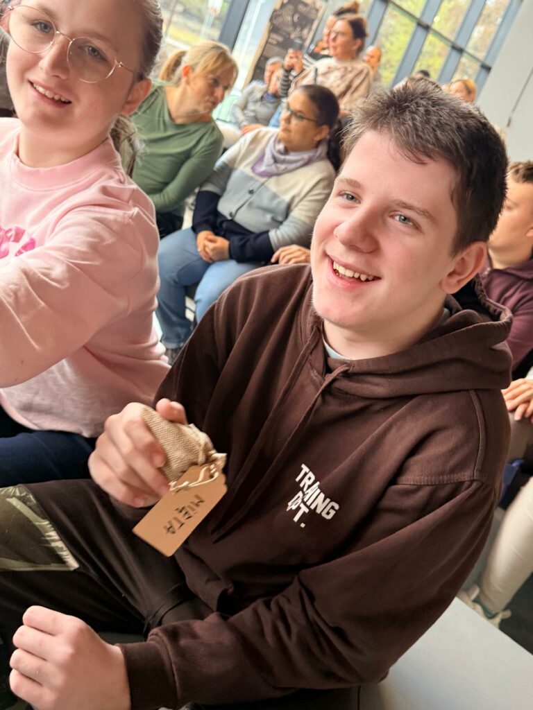 Dziewczynka w okularach i różowej bluzce oraz chłopiec w brązowej bluzie siedzą uśmiechnięci. Chłopiec trzyma w ręce woreczek z napisem "Mięta".