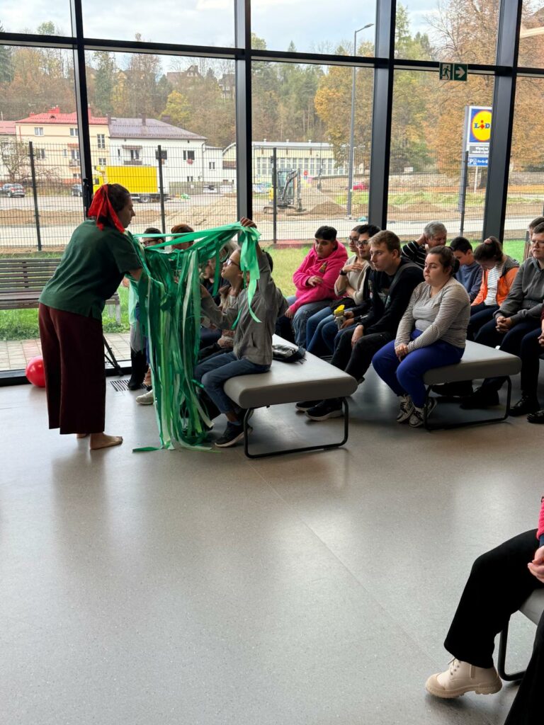 Kobieta w zielonej bluzce pokazuje grupie dzieci zielone wstążki, a uczestnicy z zainteresowaniem obserwują i dotykają materiału.