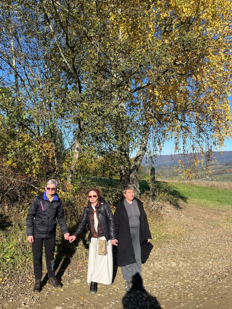 Dwie kobiety i Siostra Zakonna na tle drzew na polnej drodze.