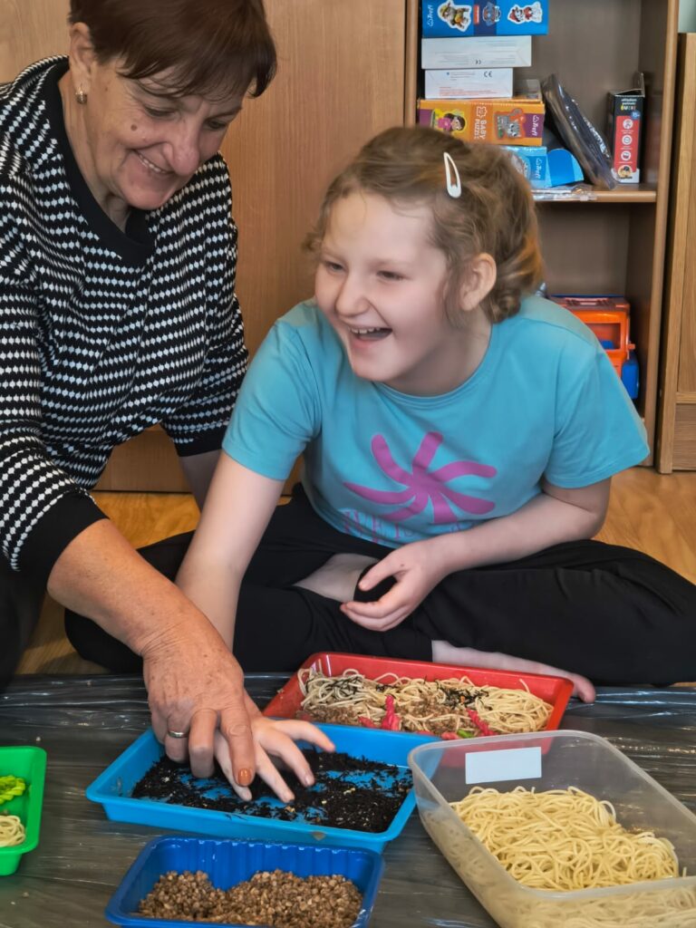 Uśmiechnięta dziewczynka razem z nauczycielką przekłada materiał sypki z jednej tacki do drugiej.