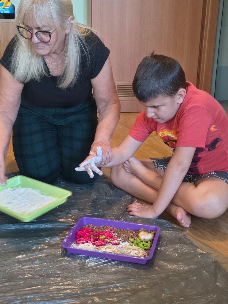 Chłopiec w czerwonej bluzce bawił się w mące z pomocą kobiety, która trzyma go za rękę.