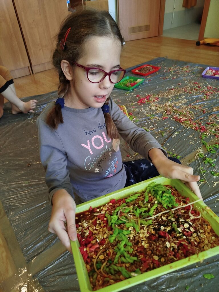 Dziewczynka w okularach trzyma w rękach miskę z różnymi makaronami, kaszą, płatkami itp. zabarwionymi na różne kolory.