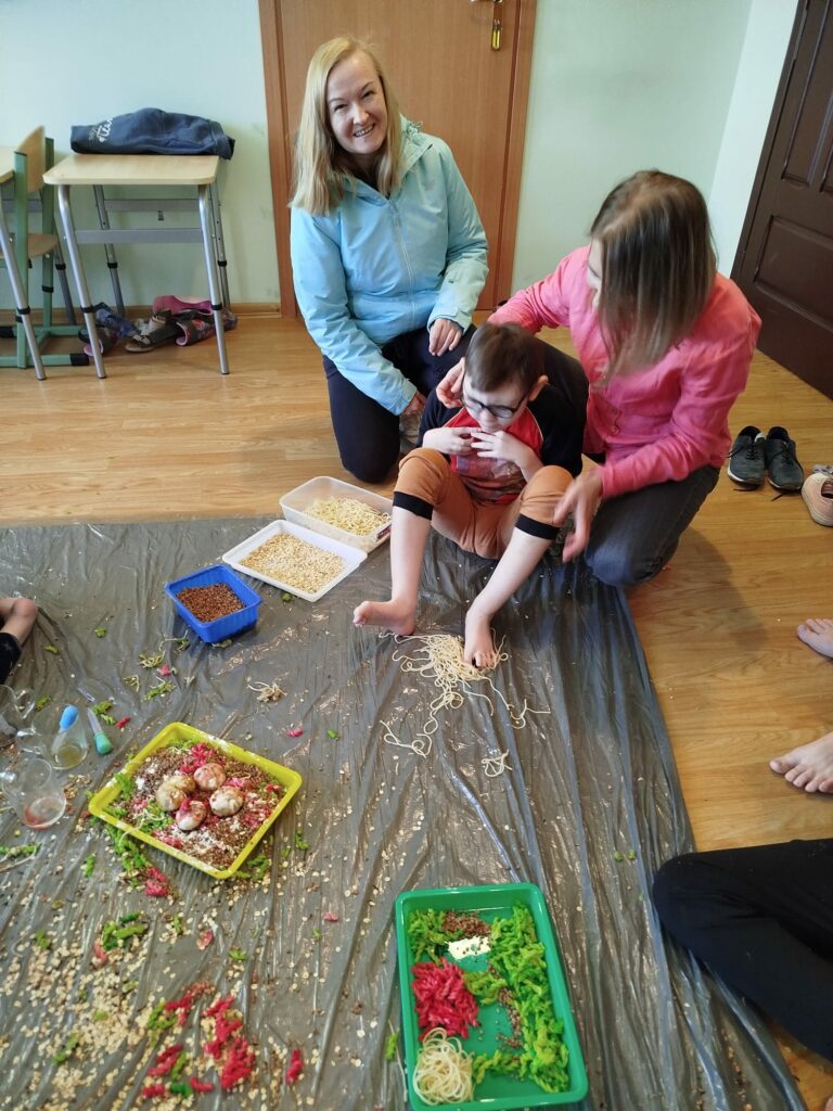 Chłopiec w okularach siedzi na podłodze na rozłożonej folii asekurowany przez kobietę kucającą za nim i dotyka gołymi stopami makaronu rozsypanego na folii. Obok klęczy druga kobieta.