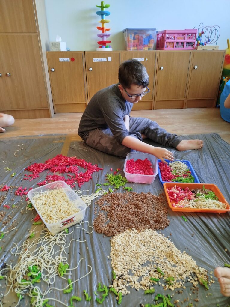 Chłopiec w okularach przekłada z pojemnika na tackę makaron zabarwiony na czerwono.