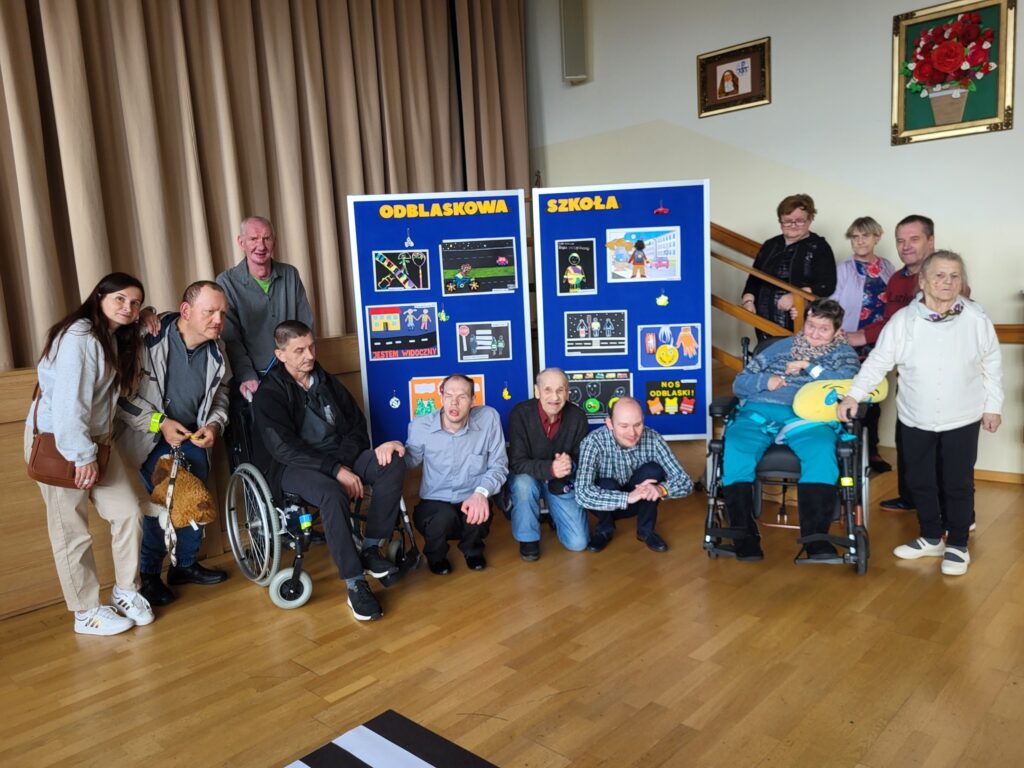 Grupa starszych osób z opiekunami pozuje do zdjęcia na tle gazetki z napisem "Odblaskowa Szkoła"