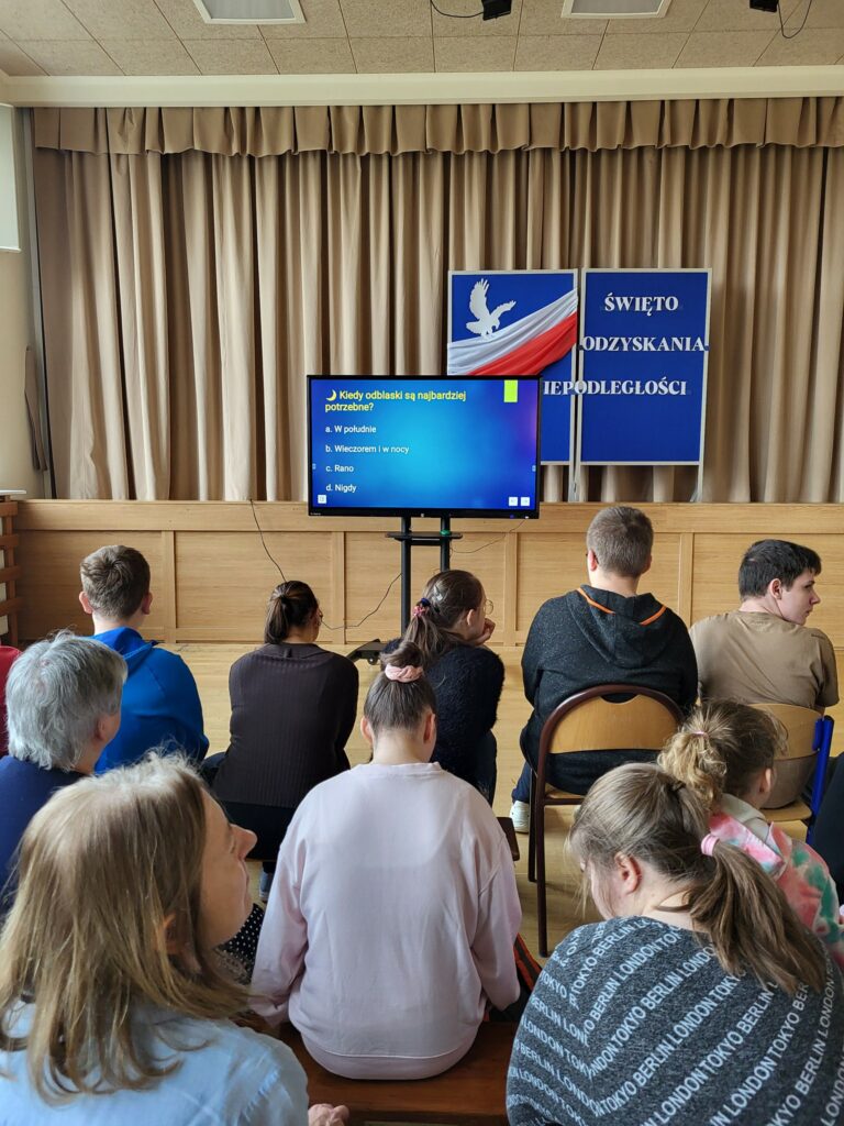 Grupa osób siedzi na sali i przygląda się pytaniu wyświetlonemu na urządzeniu multimedialnym.