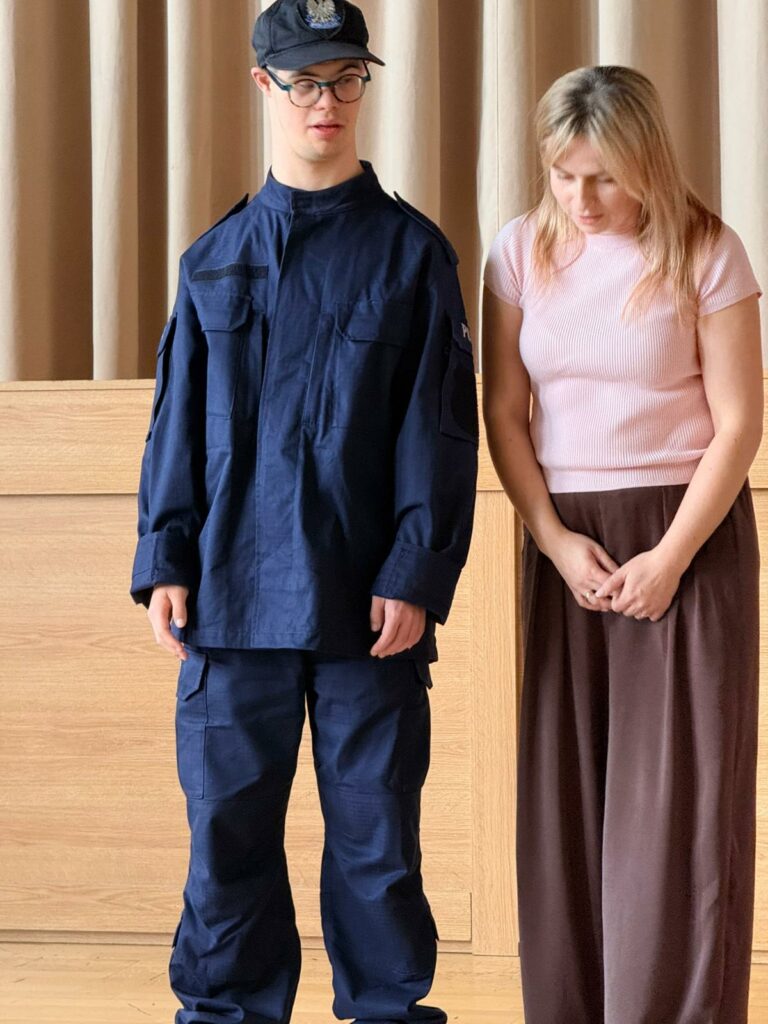 Przed sceną stoi chłopiec w policyjnym mundurze z nauczycielką.