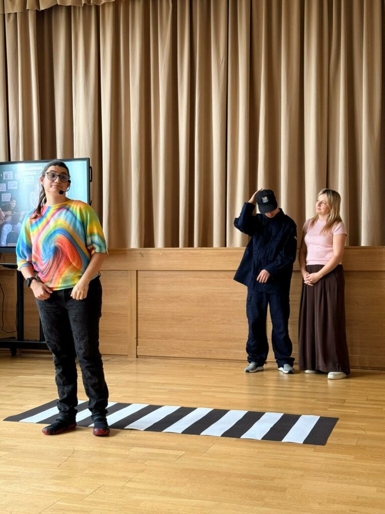 Młoda kobieta stoi na pasach za nią chłopiec w policyjnym mundurze z nauczycielką.