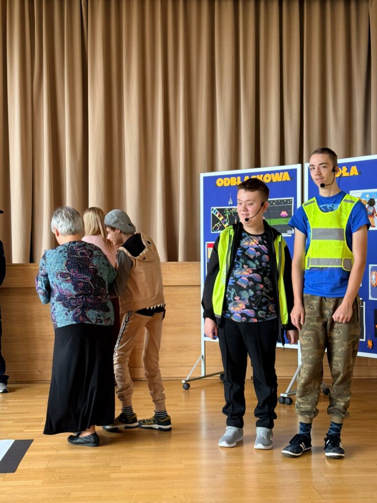 Dwaj młodzi mężczyźni w kamizelkach odblaskowych z mikrofonami stoją na tle gazetki z pracami plastycznymi. 