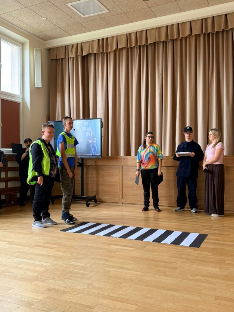 Grupa osób podczas występu. Dwóch chłopców w kamizelkach odblaskowych stoi przed przejściem dla pieszych, obok chłopiec z dziewczyną i nauczycielka.