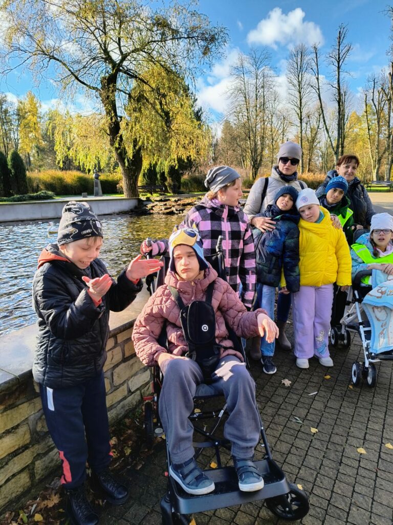 Grupa dzieci i dorosłych przy stawie w parku. Dzieci ubrane są w kolorowe kurtki, dwoje z nich siedzi na wózkach, wszyscy cieszą się spacerem w słoneczny jesienny dzień.