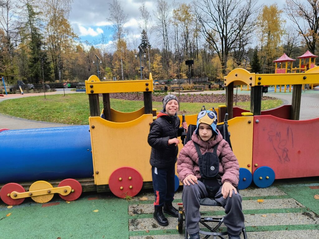 Dwóch chłopców na placu zabaw z kolorowym pociągiem. Jeden siedzi na wózku, drugi stoi obok niego i uśmiecha się szeroko. W tle widać drzewa i jesienne liście.