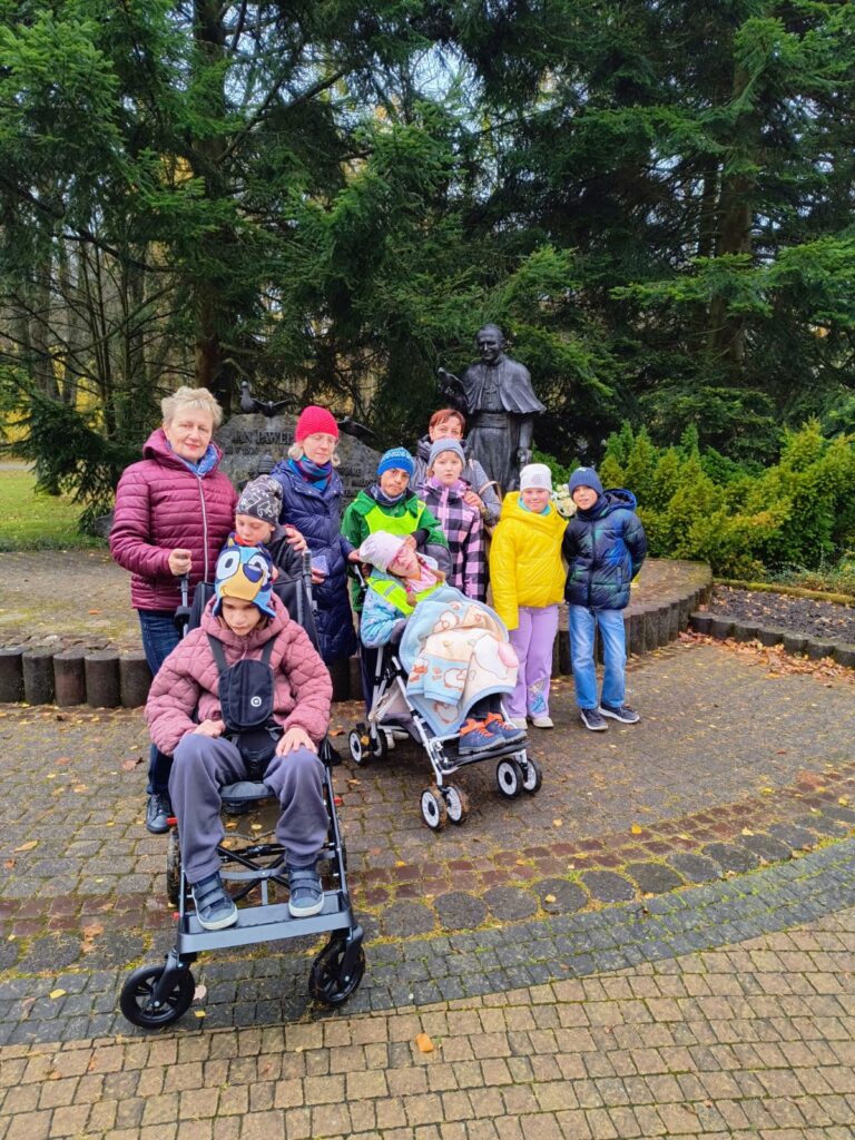 Grupa dzieci i dorosłych pozuje do zdjęcia w jesiennym parku pod pomnikiem Jana Pawła II.