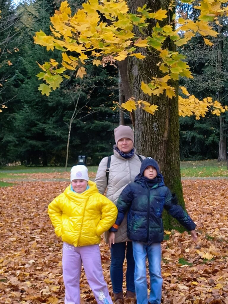 Kobieta i dwoje dzieci stoją pod drzewem z żółtymi liśćmi w jesiennym parku, otoczeni opadłymi liśćmi.