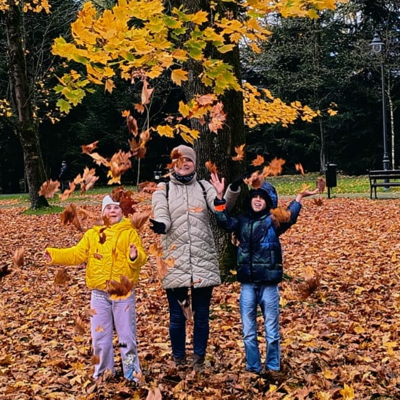 Kobieta i dwoje dzieci radośnie podrzucają jesienne liście w parku, w tle duże drzewo z żółtymi liśćmi.