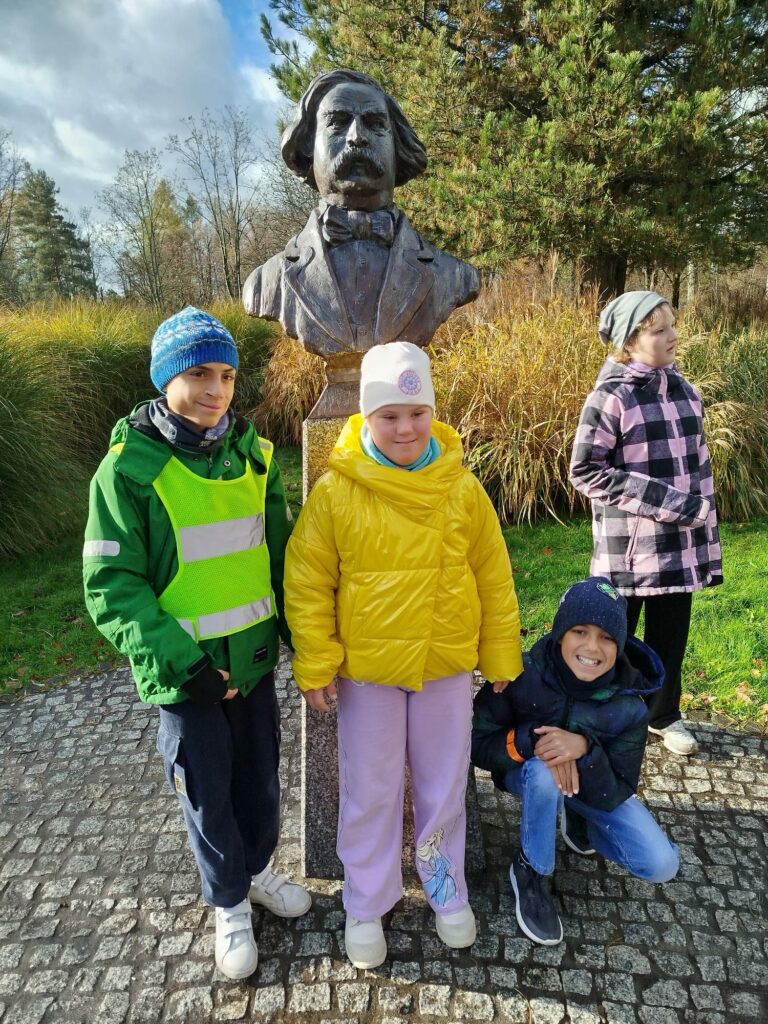 Troje dzieci pozuje przy popiersiu mężczyzny w parku, w słoneczny jesienny dzień.