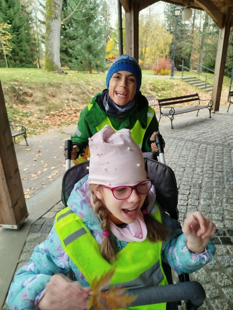 Dwoje dzieci podczas spaceru przy tężni. Dziewczynka w okularach i różowej czapce siedzi w wózku, a chłopiec w zielonej kurtce i niebieskiej czapce stoi za nią, oboje są uśmiechnięci.