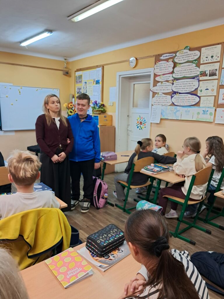 Nauczycielka wraz uczniem stoją w klasie młodszych dzieci siedzących w ławkach.