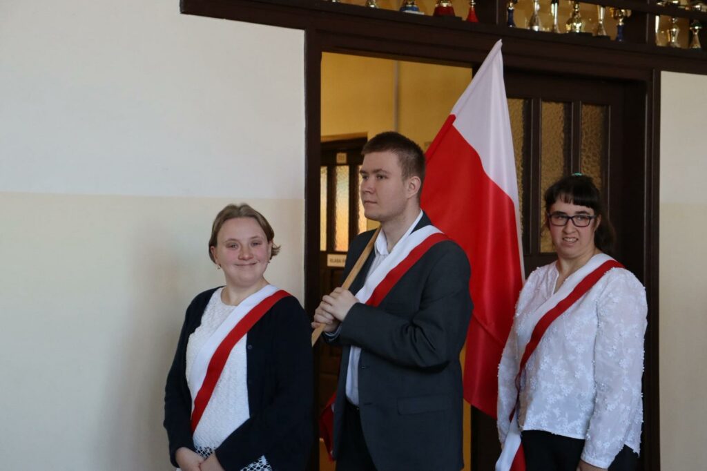 Dwie młode kobiety w strojach galowych z biało-czerwonymi szarfami i mężczyzna w garniturze z flagą Polski.