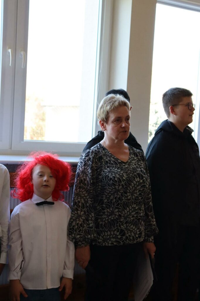 Kobieta i chłopiec w białej koszuli z muszką i czerwonej peruce na głowie stoją w pozycji na baczność.