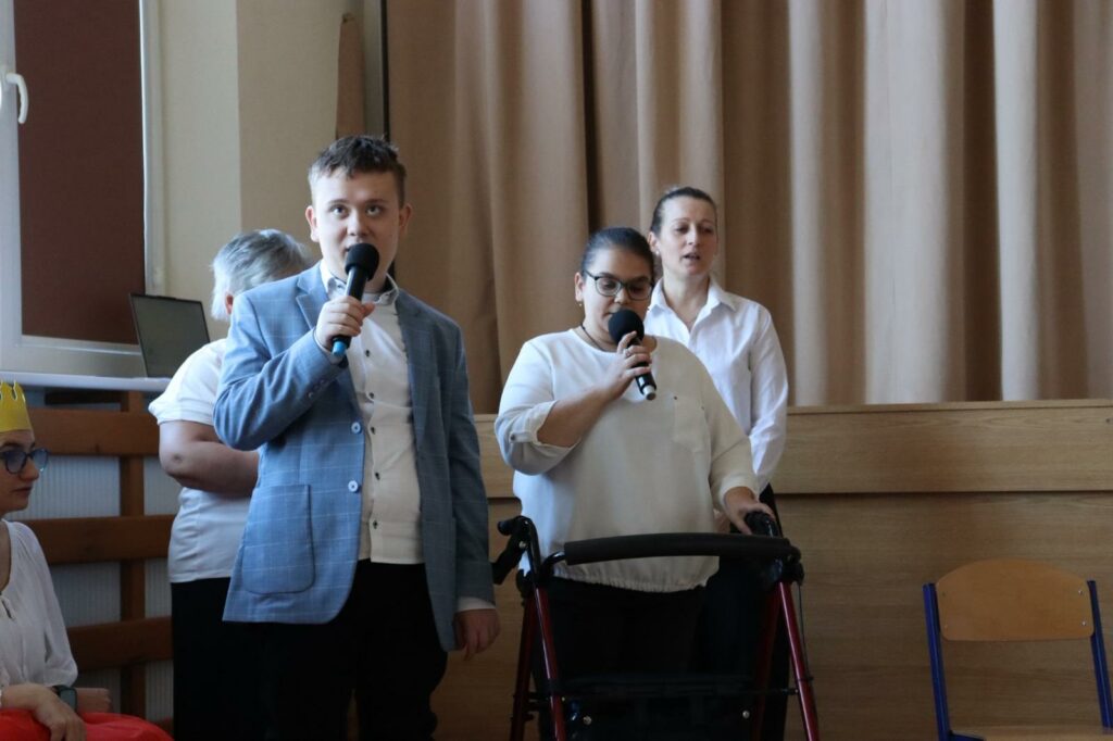 Chłopiec i dziewczyna w okularach trzymająca się chodzika, śpiewają do mikrofonów. w tle dwie kobiety w białych bluzkach.