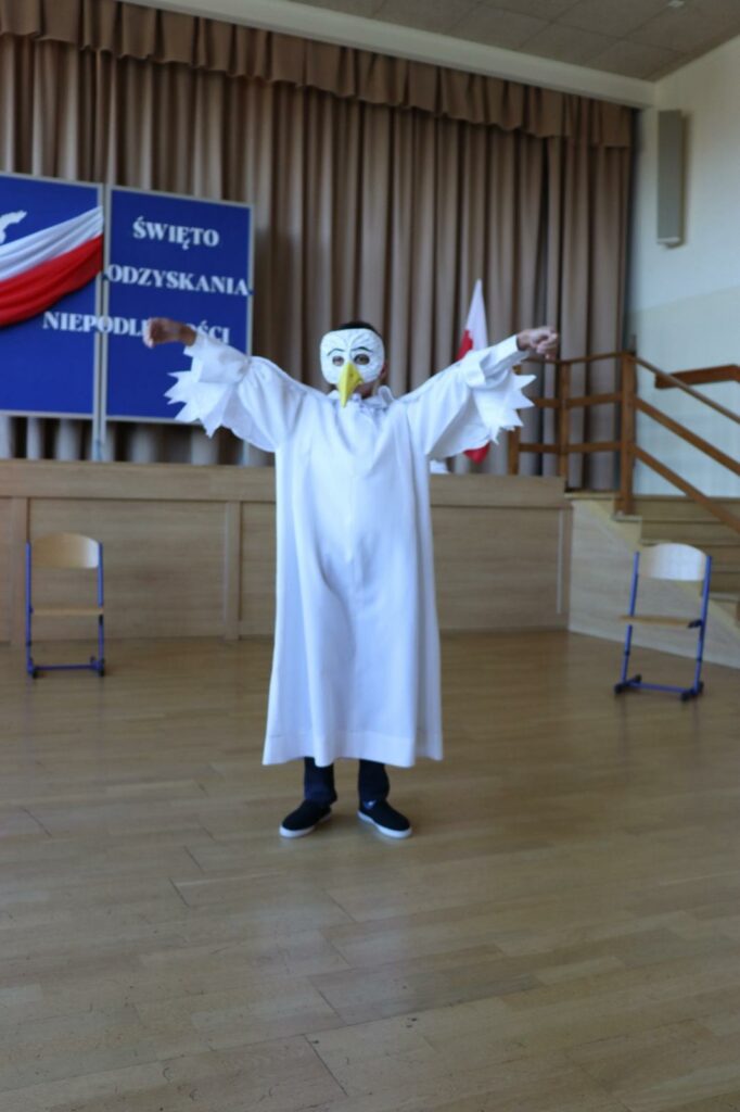 Chłopiec w stroju Białego Orła z maską na twarzy stoi z rozłożonymi rękami.
