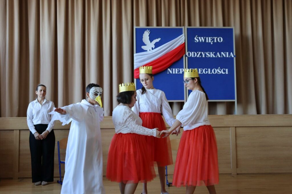 Trzy dziewczyny w białych bluzkach i czerwonych spódnicach z koronami na głowie trzymają się za ręce w kółku obok nich chłopiec w stroju Orła. za nimi na scenie gazetka z orłem, flagą Polski ni napisem "Święto Odzyskania Niepodległości".