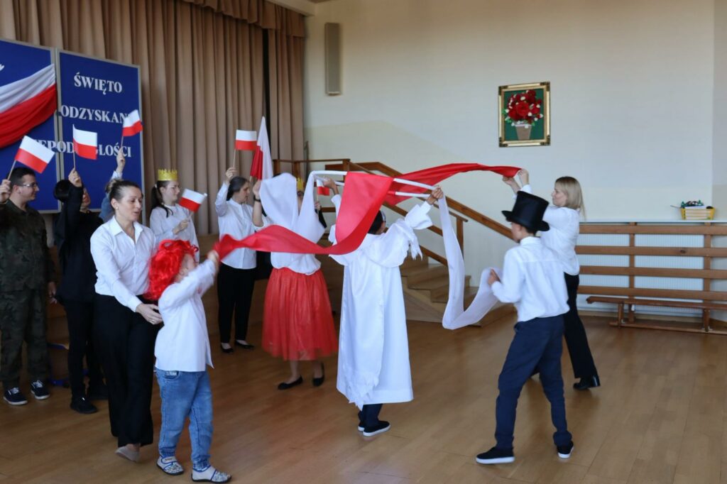 Grupa dzieci i dorosłych podczas tańca z białymi i czerwonymi szarfami. w tle pozostali uczestnicy machają biało-czerwonymi flagami.