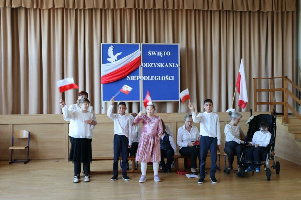 Grupa dzieci i dorosłych. Pierwszy rząd stoi trzymając w rękach flagi, drugi rząd siedzi na ławce.