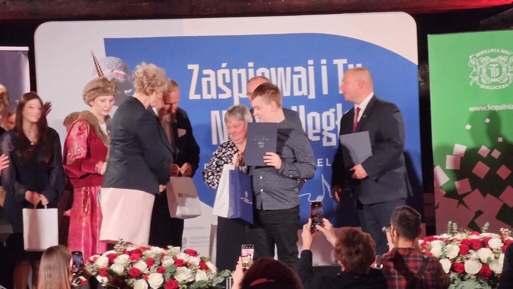 Uroczystość wręczenia nagród; grupa osób na scenie odbiera dyplomy i upominki.