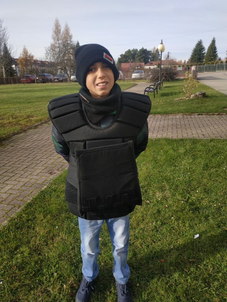 Chłopiec ma założoną policyjną kamizelkę kuloodporną stoi na zewnątrz, w tle widać trawnik, ławki i zaparkowane samochodu.