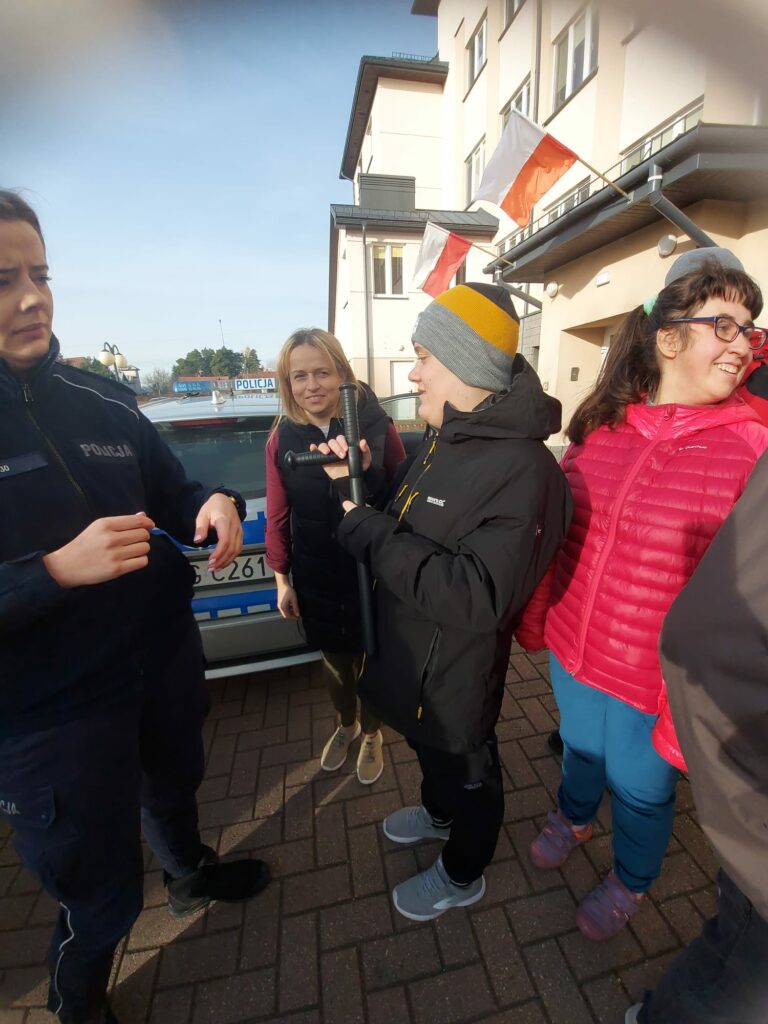 Kilka osób stoi obok policjantki przy radiowozie, jedna z osób trzyma policyjną pałkę, a reszta przygląda się z zainteresowaniem.