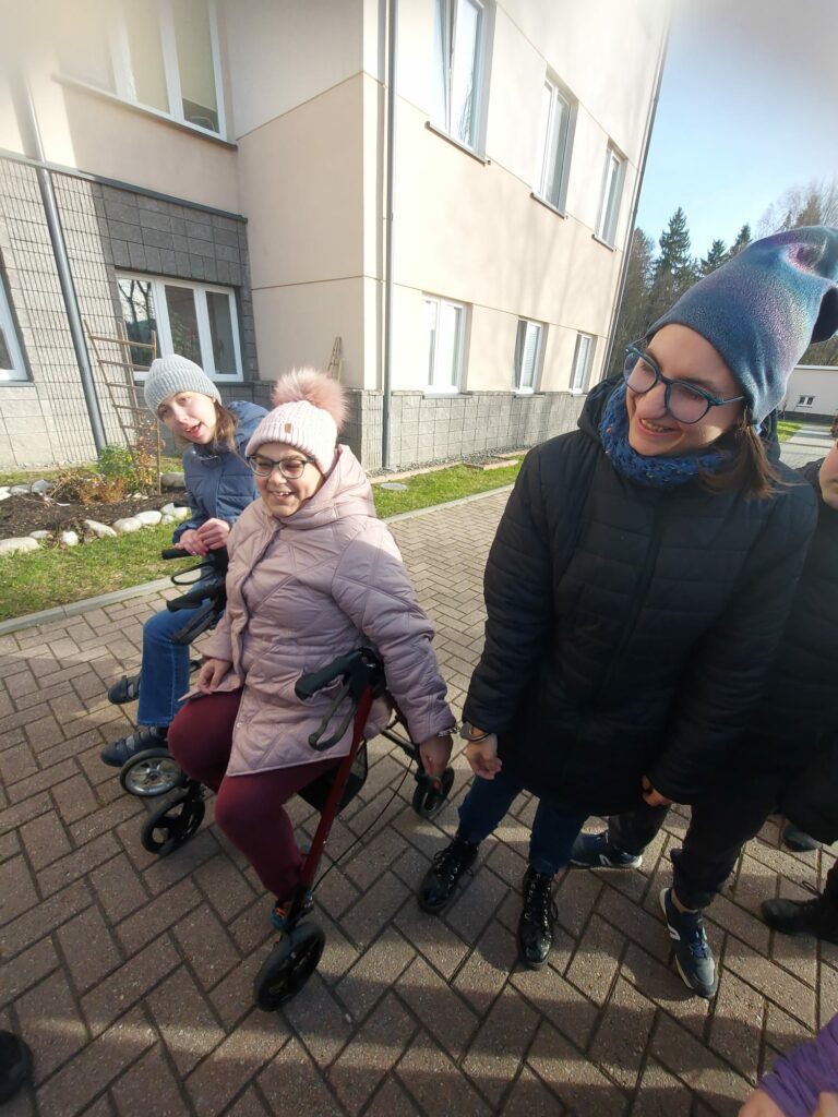 Grupa osób na dworze przed budynkiem, dwie kobiety siedzą na balkonikach, jedna z nich jest połączona kajdankami z kobietą stojącą obok.