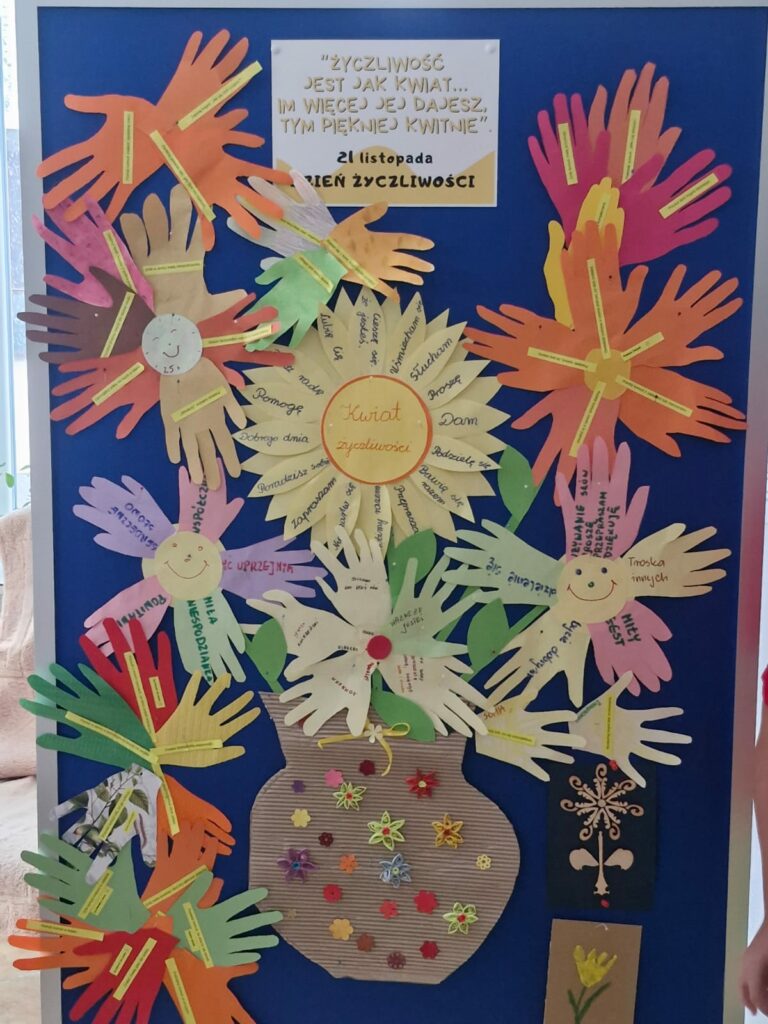 Niebieska tablica z kwiatami ułożonymi z wyciętych dłoni i napisami o życzliwości.