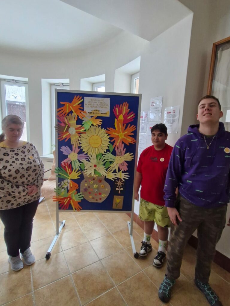 Dwóch chłopców i dziewczyna stoją po bokach tablicy z pracami plastycznymi, są to kwiaty ułożone z papierowych dłoni.