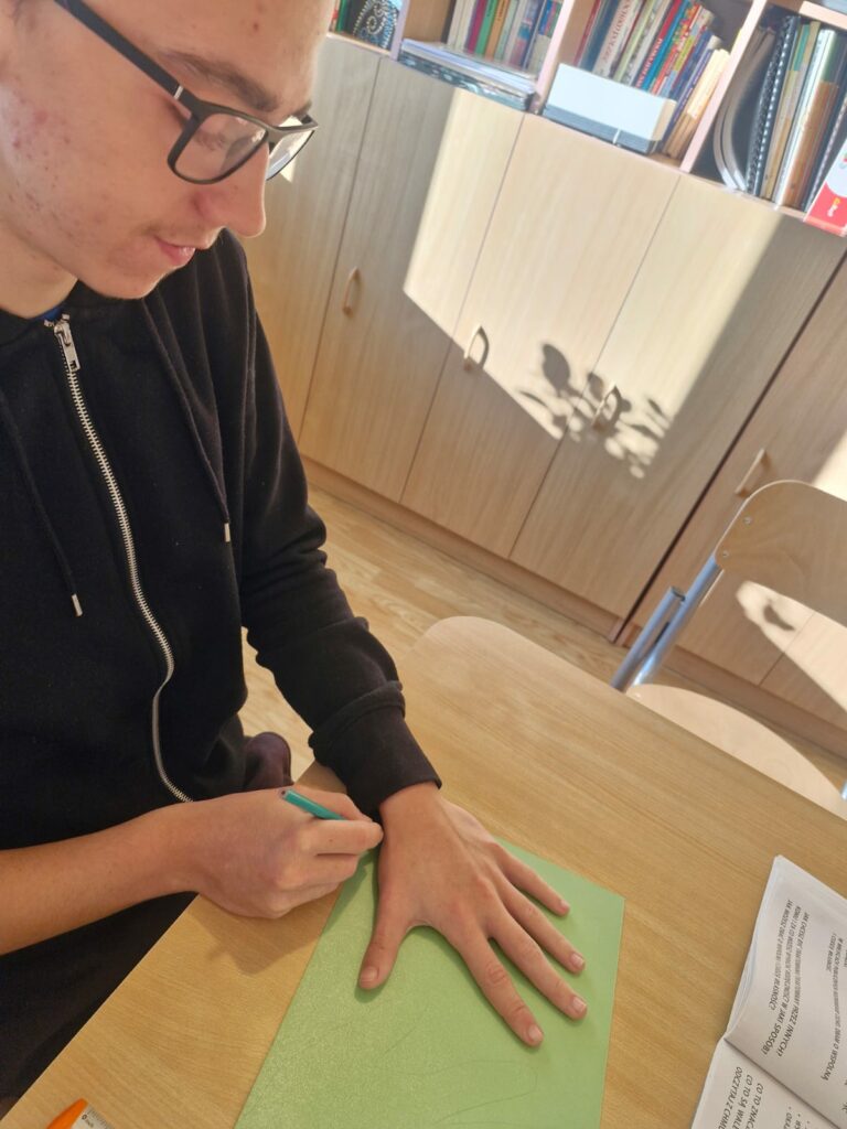 Chłopiec w okularach siedzi przy stoliku i na zielonej kartce odrysowuje swoją dłoń.