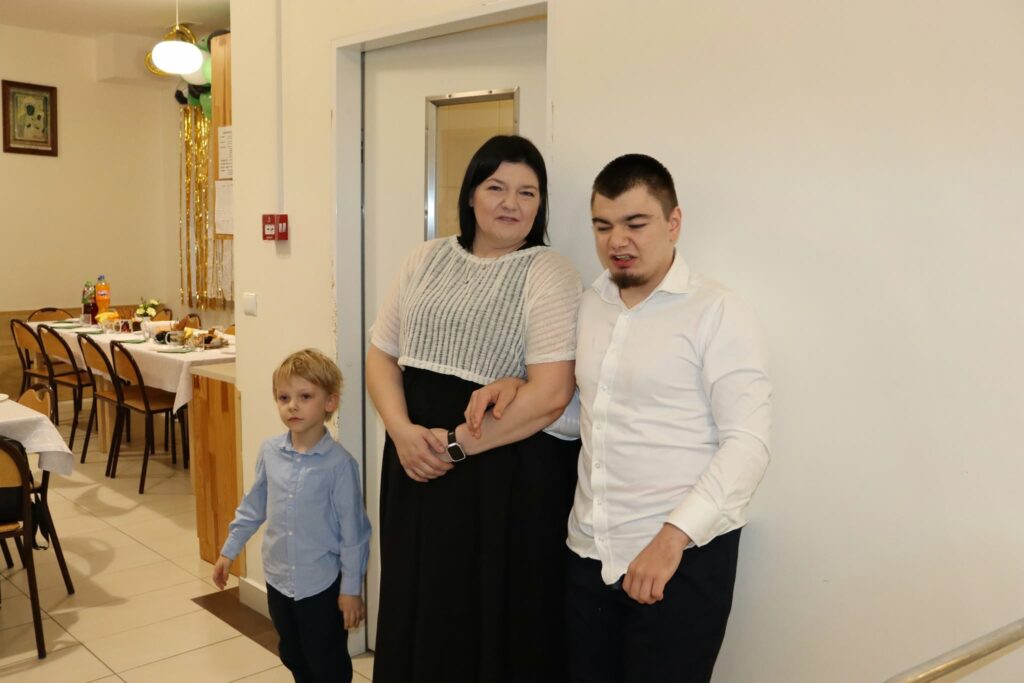 Kobieta stoi obok dwóch chłopców – starszego i młodszego – przy wejściu do sali przygotowanej na przyjęcie.