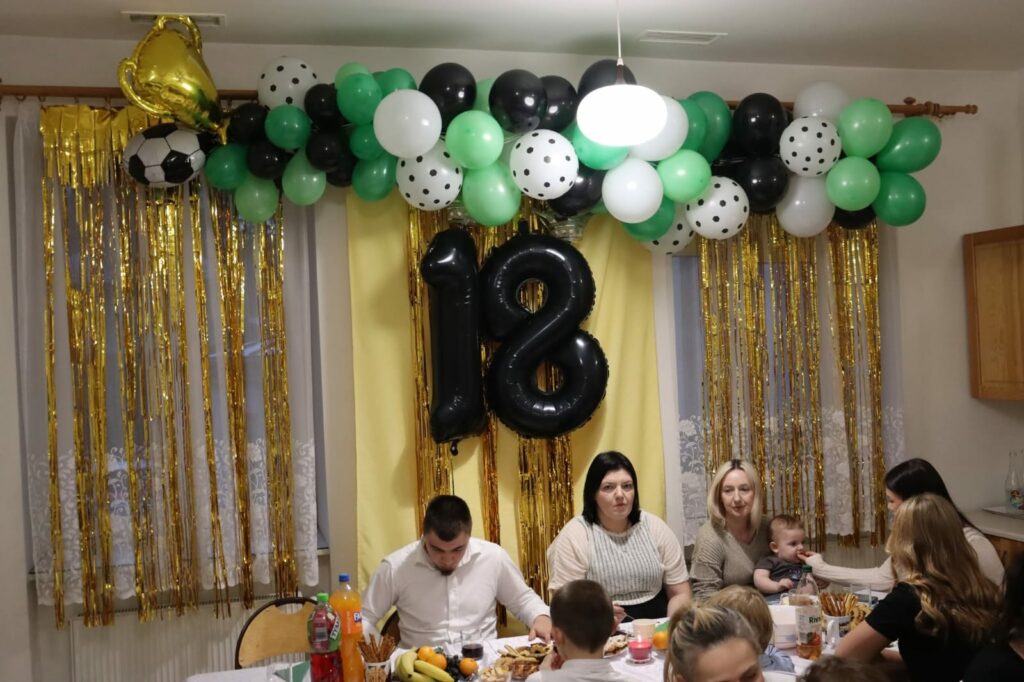 Goście siedzą przy stole pod dekoracją z balonów i złotych tasiemek, na której widnieje duży czarny napis „18”.
