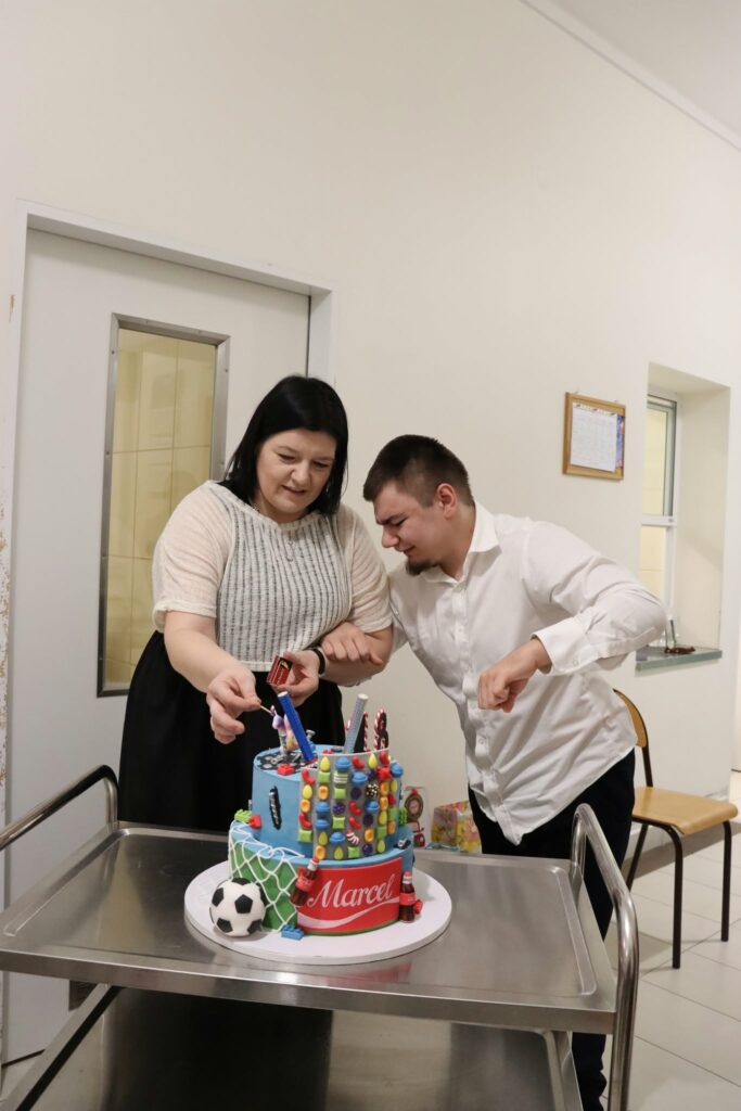 Kobieta i młody mężczyzna wspólnie zapalają świeczki na dwupiętrowym, kolorowym torcie urodzinowym z motywem piłkarskim.