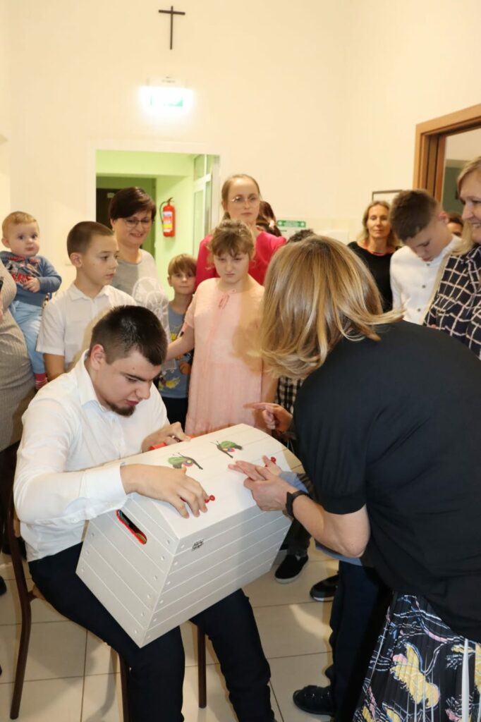 Młody mężczyzna siedzi na krześle i trzyma na kolanach pudełko z prezentem. W tle pozostali uczestnicy przyjęcia.