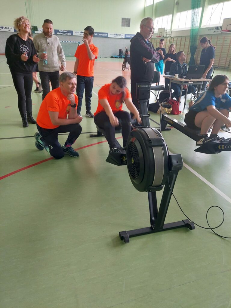Młoda zawodniczka w pomarańczowej koszulce wiosłuje na ergometrze, a obok klęczy instruktor, w tle widać obserwującą grupę.