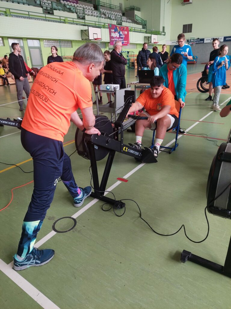 Osoba w pomarańczowej koszulce ćwiczy na ergometrze, wspierana przez instruktora stojącego obok.