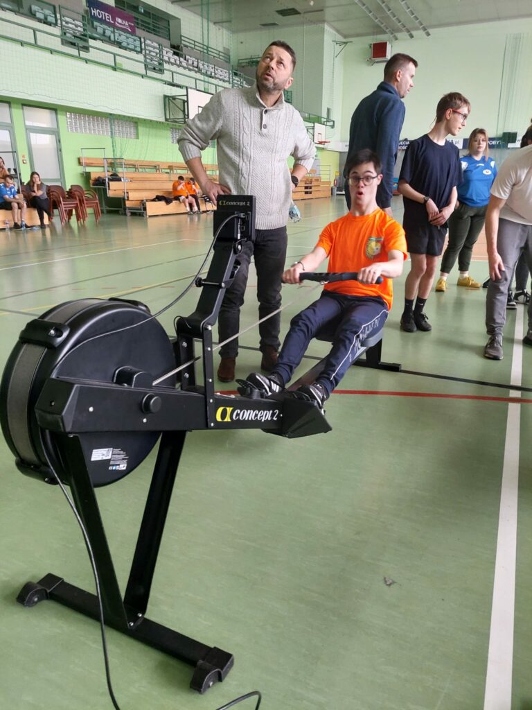 Uczestnik w pomarańczowej koszulce intensywnie ćwiczy na ergometrze, a stojący obok mężczyzna uważnie obserwuje jego wyniki.