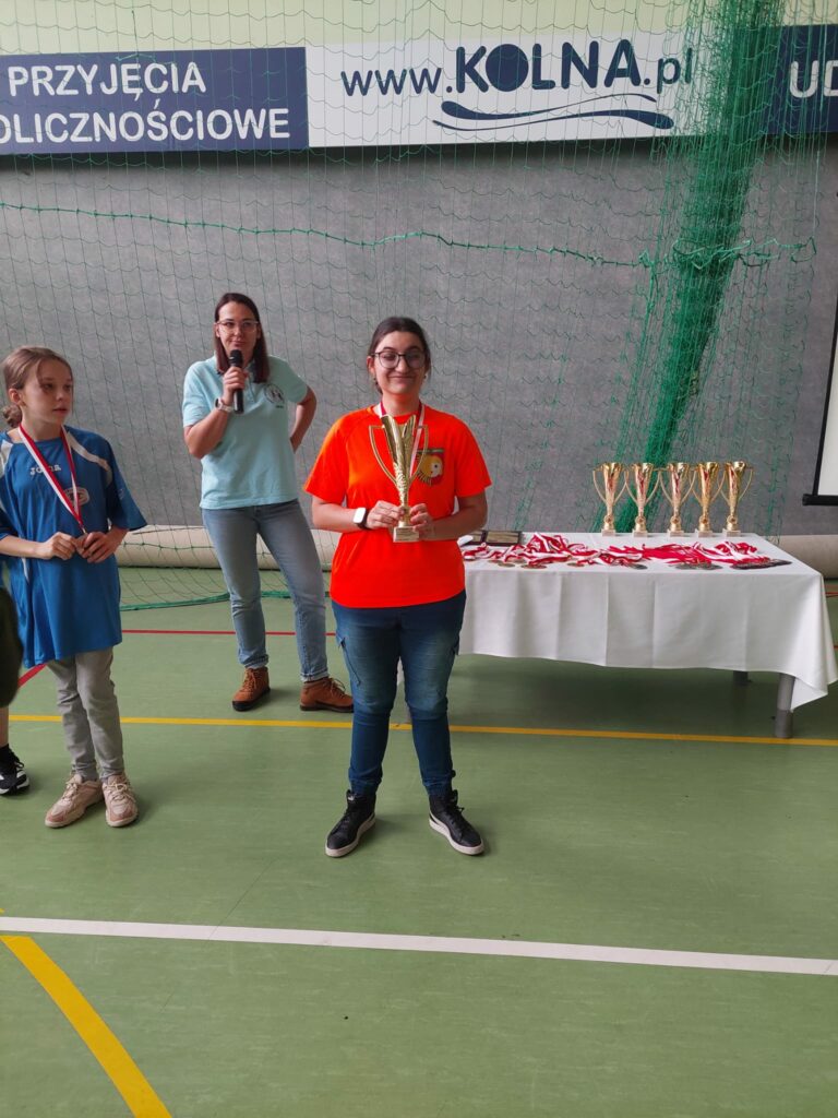 Młoda kobieta w pomarańczowej koszulce trzyma w rękach puchar i uśmiecha się.