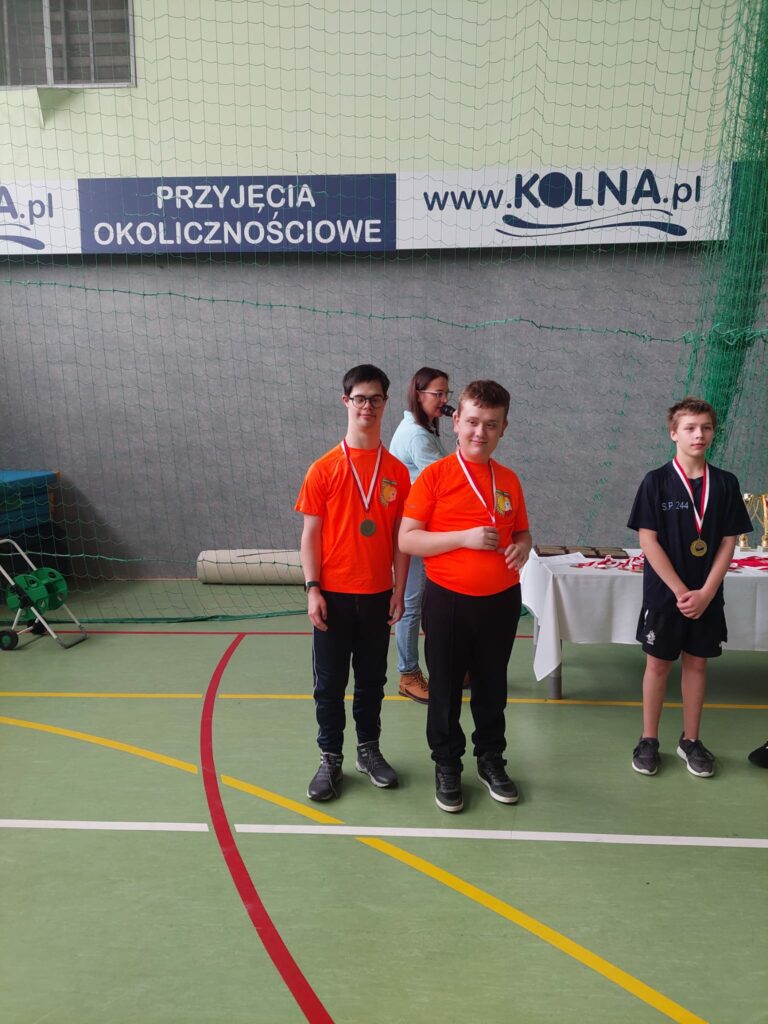 Dwóch chłopców w pomarańczowych koszulkach i jeden w czarnej z medalami na szyi, pozują do zdjęcia.