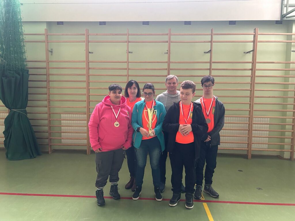 Grupa młodzieży z opiekunami podczas zawodów z medalami i pucharem pozuje do zdjęcia. W tle widać drabinki.