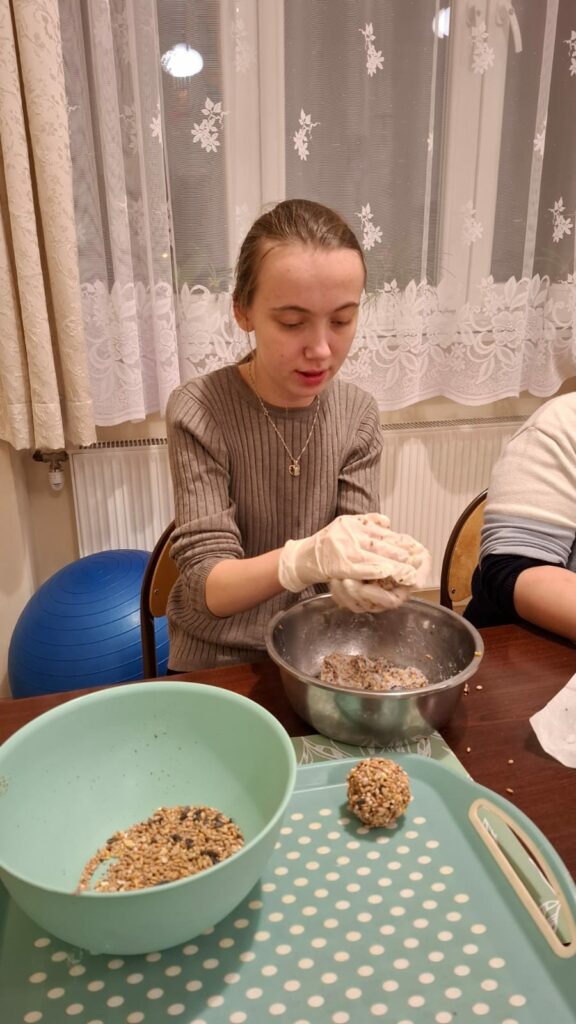 Młoda kobieta w brązowym swetrze i jednorazowych rękawiczkach siedzi przy stole, Przed nią stoi miska. Kobieta formuje w rękach kulę z masy.