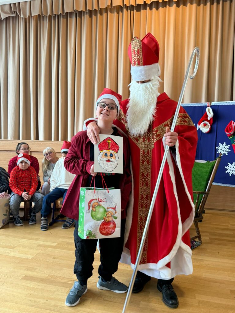 Chłopiec w świątecznej czapce stoi obok św. Mikołaja i trzyma torbę z prezentem oraz własnoręcznie wykonany rysunek Mikołaja.