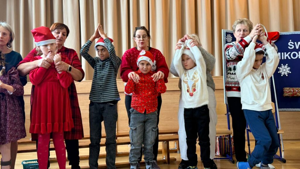 Grupa dzieci i dorosłych w świątecznych strojach stoi na scenie; dorośli pomagają dzieciom wykonywać gesty podczas wspólnego występu.